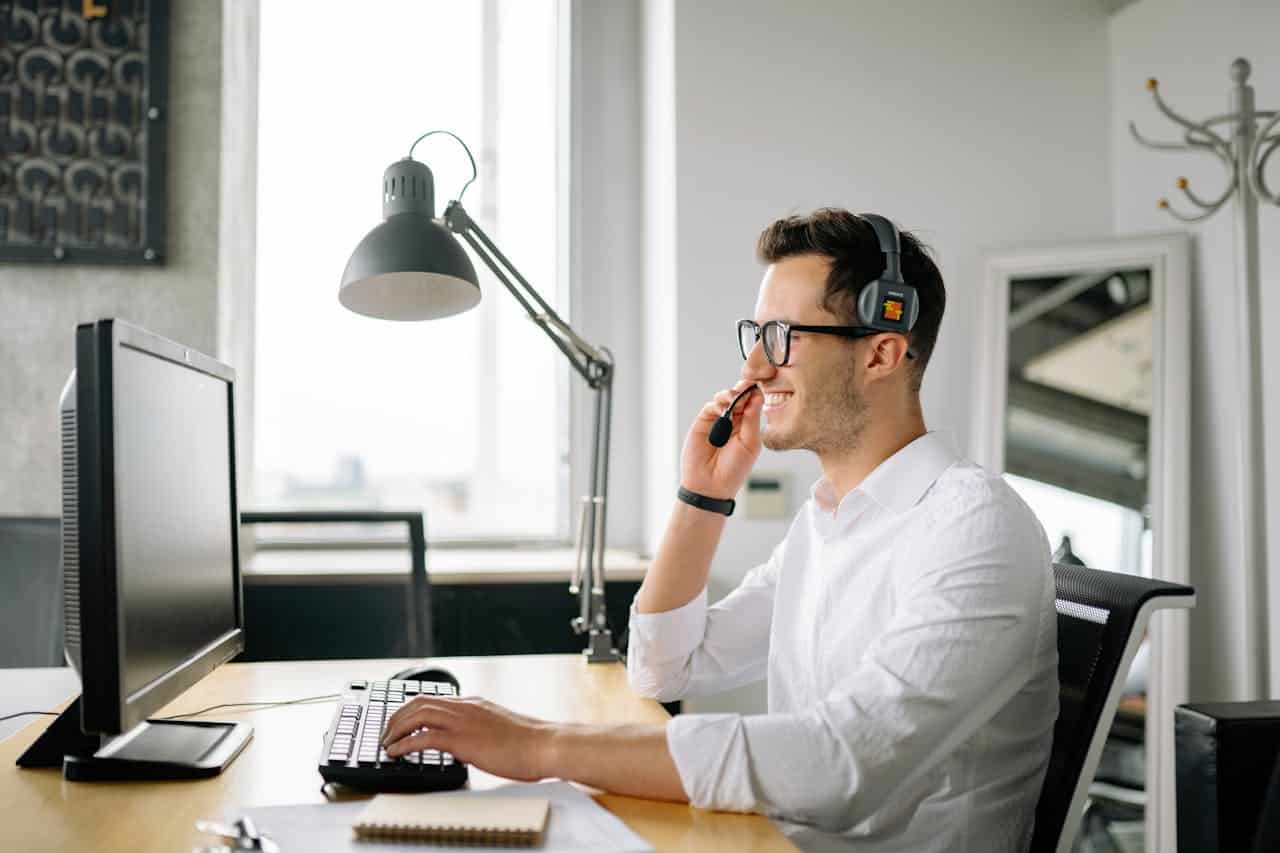 A man wearing glasses sits at a desk with a computer and a phone, focused on his work.