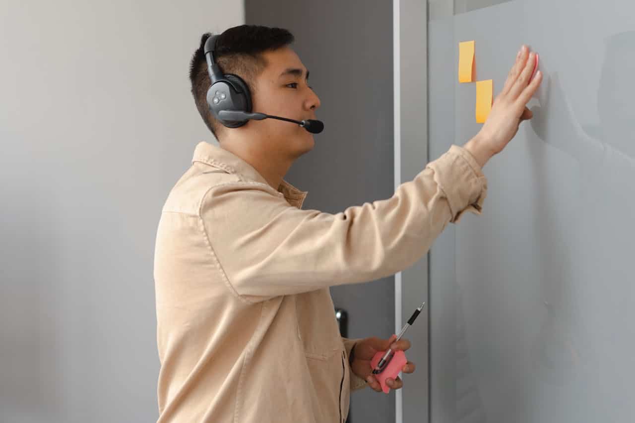 A man with headphones holds a sticky note on a wall, focused on his task.